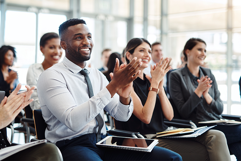 Group of sitting employees smile and clap as they listen to a presentation in front of them 